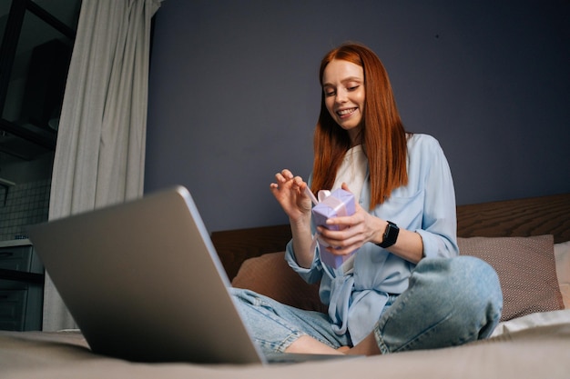 Lage hoekopname van een vrolijke roodharige jonge vrouw die huiskleding draagt die een geschenkdoos met cadeau draagt terwijl ze een videogesprek voert op een laptop die thuis op bed zit in een lichte slaapkamer.