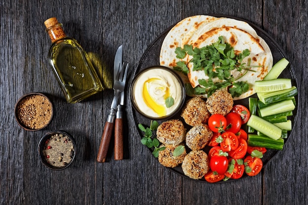 Lamb meatballs served with flatbread, fresh cucumber, tomatoes and hummus on a black plate on a wooden table, flat lay