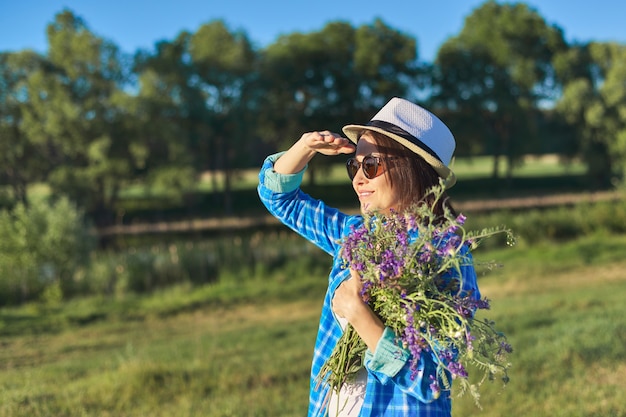 Land portret van een volwassen mooie vrouw in hoed met boeket van wilde bloemen. Groen natuurlijk landschap, lente-zomerweiden, vrouw kijkt weg, kopieer ruimte