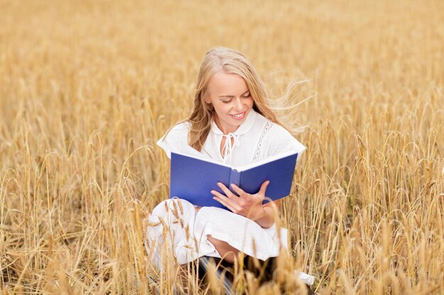 land, zomervakantie, literatuur en mensenconcept - lachende jonge vrouw in witte jurk leesboek over graanveld