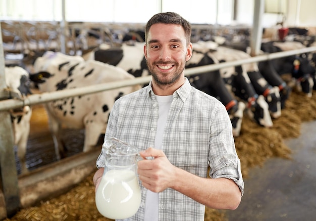 landbouw industrie, landbouw, mensen en veehouderij concept - gelukkige glimlachende jonge man of boer met koeien melk in kruik bij koeienstal op melkveehouderij