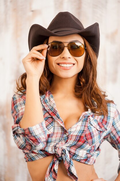 Landelijk meisje. Mooie jonge cowgirl die haar bril aanpast en naar de camera glimlacht terwijl ze tegen de houten achtergrond staat