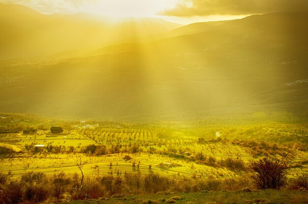 Landelijk zonsonderganglandschap met groene heuvels, dramatische lucht, bergen en zonnestralen