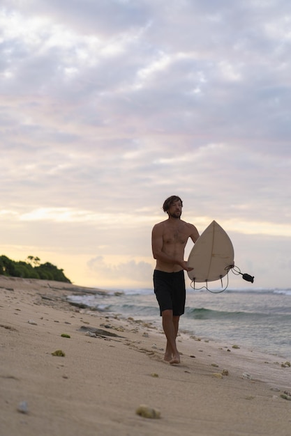 Immagine del paesaggio del surfista maschio impegnato a camminare sulla spiaggia all'alba mentre trasportava la sua tavola da surf sotto il braccio con le onde dell'oceano che si infrangono sullo sfondo. giovane surfista maschio bello sull'oceano