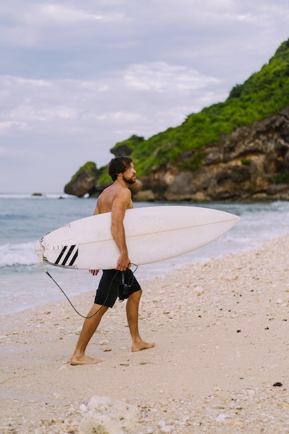 海の波を背景にサーフボードを腕に抱えて日の出のビーチを歩くのに忙しい男性サーファーの風景画像。海の上の若いハンサムな男性サーファー