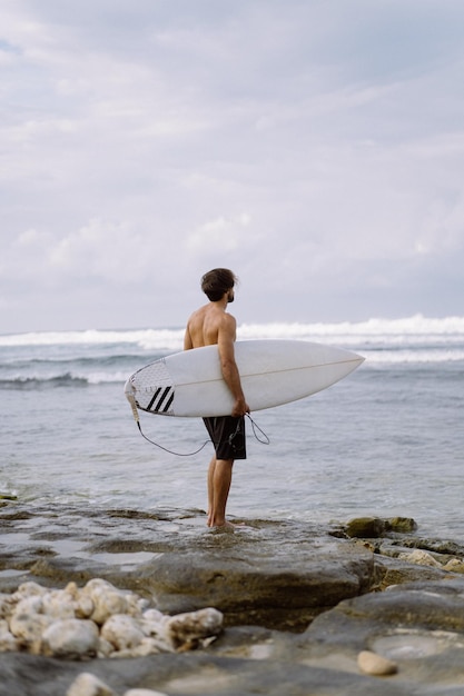 Ландшафтное изображение мужчины-серфера, занятого прогулкой по пляжу на рассвете, неся свою доску для серфинга под мышкой на фоне океанских волн, разбивающихся на заднем плане. Молодой красивый мужчина-серфер на берегу океана