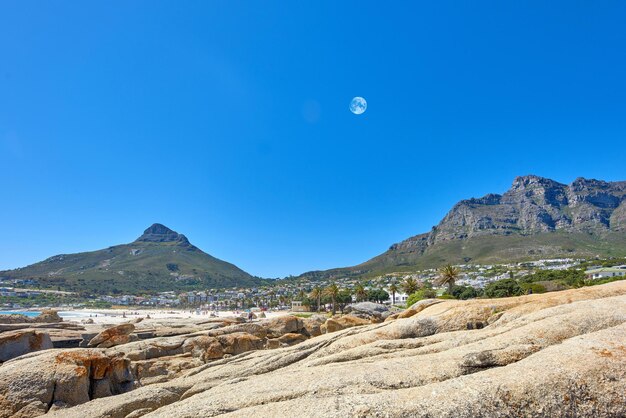 복사 공간이 있는 푸른 하늘에 산과 달의 풍경 해안선 또는 베이 지역 근처 산꼭대기의 아름다운 바위 노두 남아프리카 케이프 타운의 데블스 피크와 테이블 마운틴의 전망