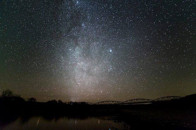 Il paesaggio della sponda del fiume dei ciottoli, gli alberi sull'orizzonte, le stelle luminose e la galassia della via lattea in cielo scuro hanno riflesso in acqua calma. bellezza del concetto di natura.