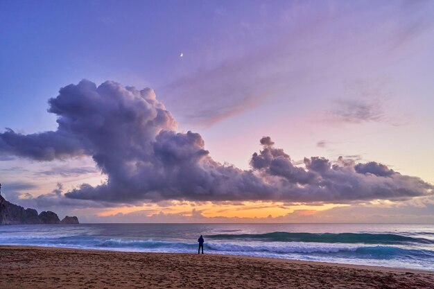 Пейзаж живописных идиллических мирных обоев спокойного неба с пушистыми облаками и силуэтом человека, стоящего в одиночестве на морском побережье и песчаном пляже