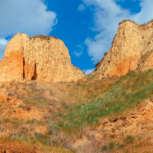 Landscape view clay cape cliffs. Green grass, lawns grown on slopes of clay mountains against blue sky. Hills of mountain and slopes of cape are overgrown with half grass and plants. Clay wall Square.