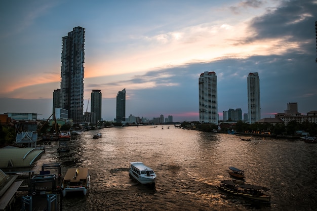 Landscape view of sunset at Chaophraya river with a view of boats and modern building along the riverside.