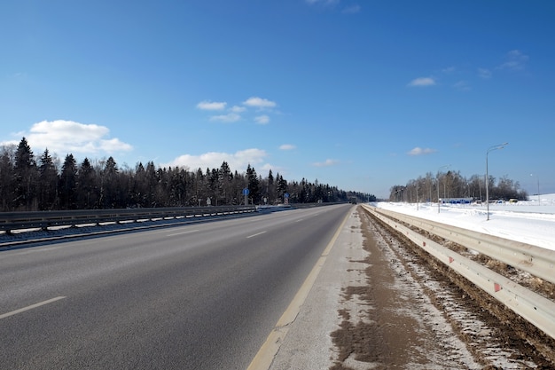 Landscape with long straight suburban highway with metal a fence on the sides and forest under clear blue sky on bright sunny winter day