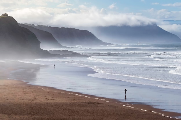 Landschap van een strand gewassen door de golven in blauwe tinten, mist en wolken op de achtergrond en een persoon die langs de kust van het strand loopt. vizcaya, baskenland. spanje.