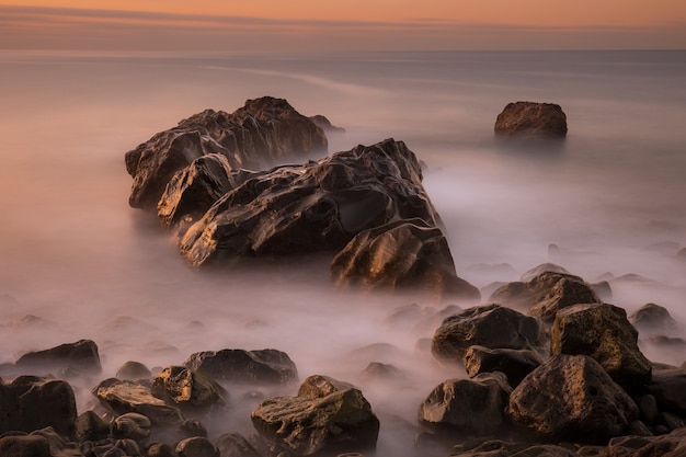 Lang blootstellingszeegezicht bij zonsopgang met rotsen dichtbij de kust met een schittering van de zon op natte stenen en melkachtig vaag water
