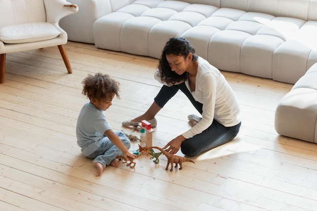 Lang geleden Glimlachende duizendjarige Afrikaanse moeder, oudere zus of babysitter spelen met kleine geïnteresseerde zwarte jongen op verwarmde houten vloer met speelgoeddinosaurusfiguren en kubussen die een fantasiewereld creëren