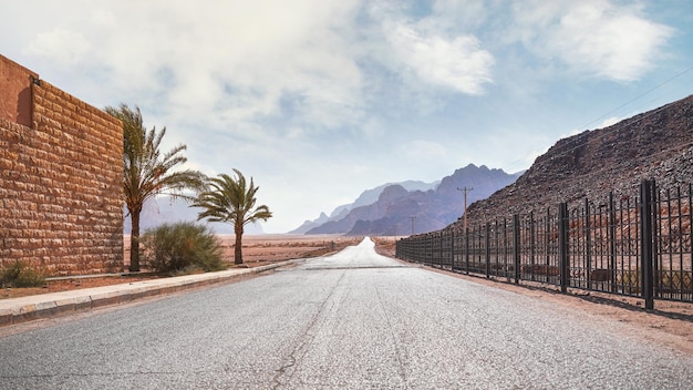 Lange rechte weg met woestijnheuvels in de verte, bakstenen muur en ijzeren hek aan de zijkanten. Landschap bij de ingang van het beschermde gebied van Wadi Rum in Jordanië