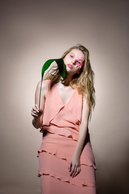 Lange stijlvolle jurk. Blond model draagt een lange stijlvolle jurk die poseert met groen blad en bloemen in de buurt van het gezicht