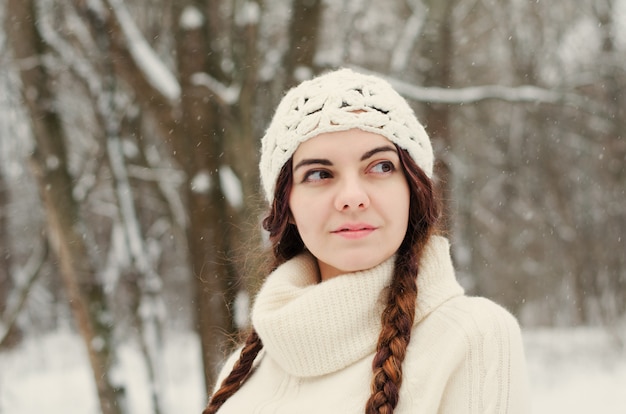 Langharig donkerbruin meisje dat in een de winterpark loopt en met sneeuw speelt, witte sweater en een met de hand gemaakte witte hoed draagt