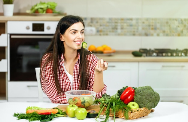 Langharig kaukasisch meisje dat geniet van een gezonde gemengde salade met een cool geruit rood hemd binnenshuis geschoten in een lichte grote keuken met een tafel vol groenten