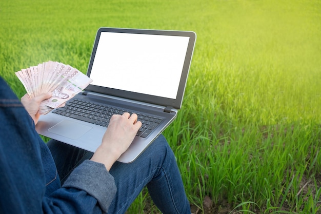 Laptop computer blank screen and banknote money are held by farmer at green rice farm