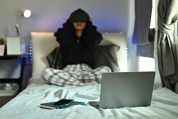 ラップトップ コンピューターとベッドの上の本、背景に座っている仮想現実ゴーグルを装着した 10 代の女性