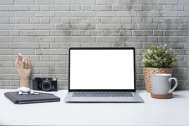 Laptop computer coffee cup camera and houseplant on white table against brick wall Blank screen for your advertise text