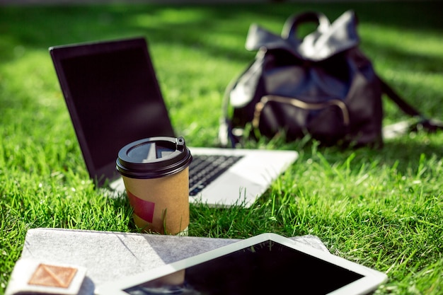 屋外公園のコーヒーカップ、バッグ、タブレットと緑の芝生の上のラップトップコンピューター。スペースをコピーします。静物