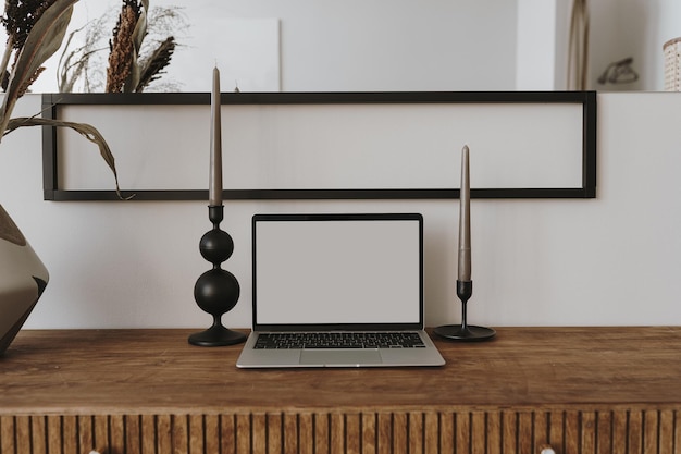 Laptop computer with blank screen on table with candles flowers bouquet Aesthetic influencer boho styled home workspace interior design template with mockup copy space Online store blog branding