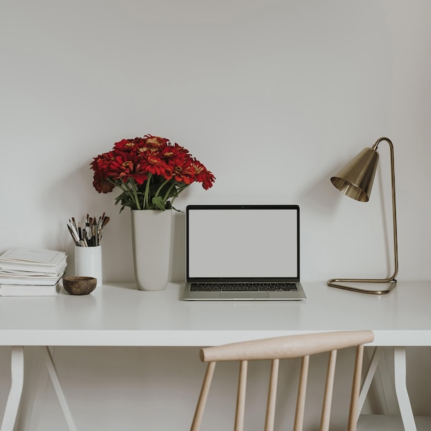 Laptop computer with blank screen on table with red gerber flowers bouquet Aesthetic influencer styled workspace interior design template with mockup copy space