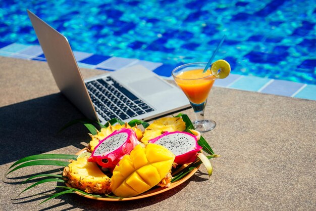 Laptop computer with blows of fresh tropical fruits papaya pineapple dragon fruit by swimming pool remote work freelance and frilansing