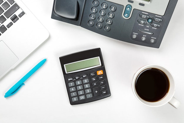 Laptop computer with cup of coffee, blue pen and modern IP phone and calculator on white surface. T