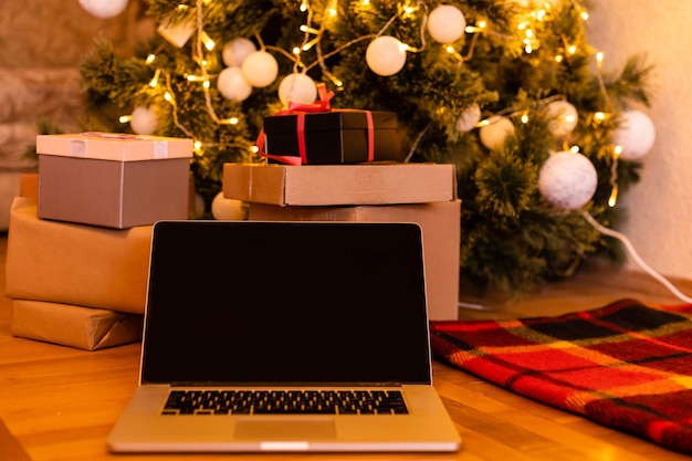 흰색 빈 모의 화면이 있는 노트북 컴퓨터에는 선물 선물, 산타 하우스 배경에 장식된 크리스마스 트리가 있습니다. 전자상거래 웹사이트 온라인 쇼핑 배달 광고.
