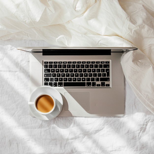 Laptop and cup of coffee on white bed with a  blanket. Work at home concept. Morning light. Lifestyle Concept