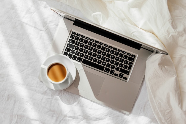 Laptop and cup of coffee on white bed with a  blanket. Work at home concept. Morning light. Lifestyle Concept