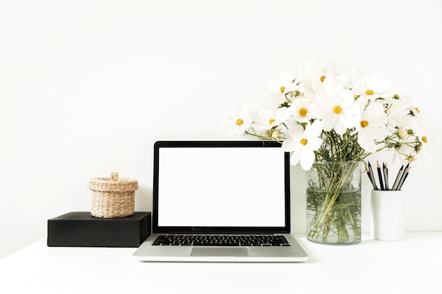 Laptop die zich op witte lijst tegen witte muur met madeliefjes in vaas, zwarte doos, rieten mand bevindt.