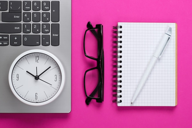 Laptop en wekker, bril, notitieboekje op roze oppervlak. De tijd loopt weg. Het concept van urgente deadlines op het werk en verplichtingen. Bovenaanzicht