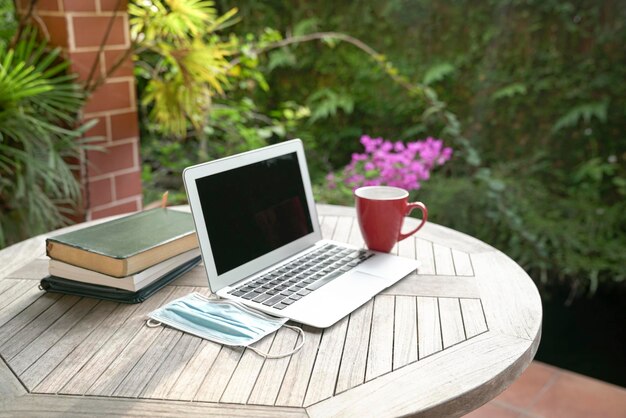 Laptop face mask books and red cup on top of table next to garden