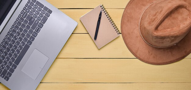 Laptop, felt hat, notebook and pen on a yellow wooden table