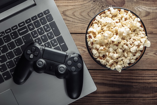 Laptop, gamepad and bowl of popcorn on wooden table