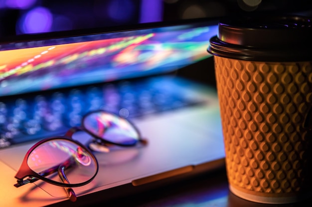 A laptop half closed in the dark with colourful glow cup of coffee and glasses