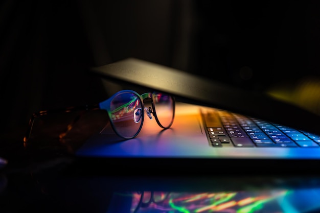 A laptop half closed in the dark with colourful glow and glasses