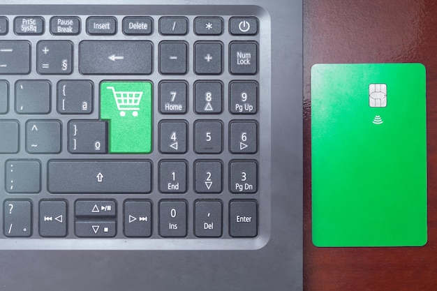 Laptop keyboard with shopping cart icon on green key and credit\
card beside.