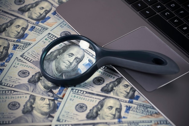 Laptop and magnifying glass on us dollar bills Benjamin Franklin portrait Online business