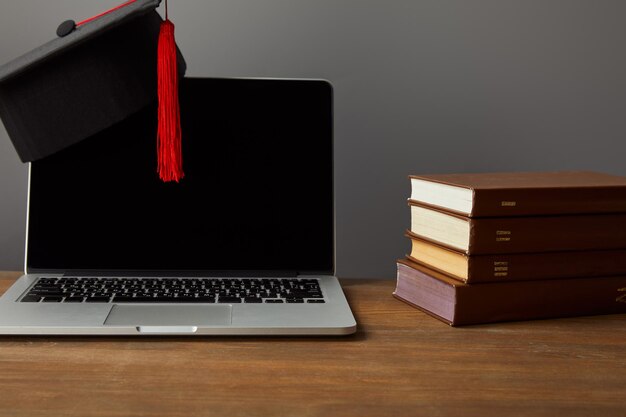 Foto laptop met blanco schermboeken en academische dop met rode kwast op houten oppervlak geïsoleerd op grijs