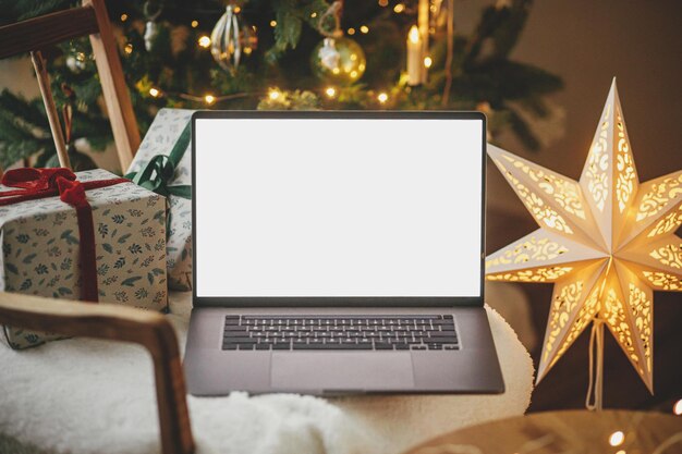 Laptop met leeg scherm en stijlvolle kerstcadeaus op moderne stoel op de achtergrond van feestelijk versierde boom gouden lichten en ster in kamer Kerstinkopen online en verkoop Laptop mock up