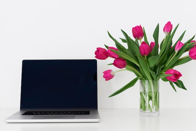 Laptop met leeg zwart scherm en boeket roze tulpenbloemen in een transparante vaas op een tafel