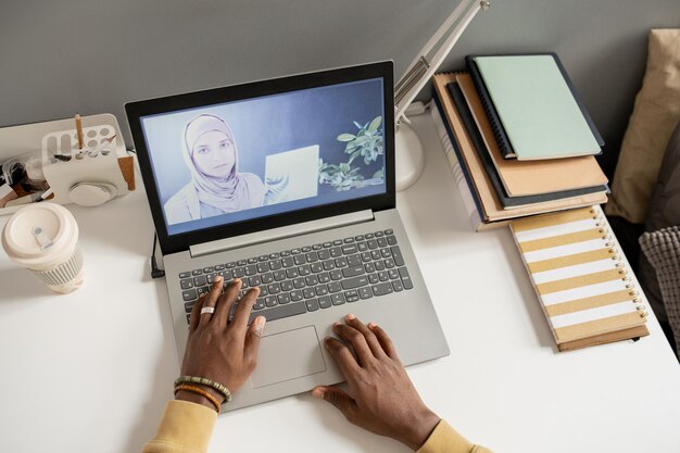 Laptop op bureau met leraar in hijab op het scherm tijdens online les en handen van afrikaanse mannelijke student over toetsenbord