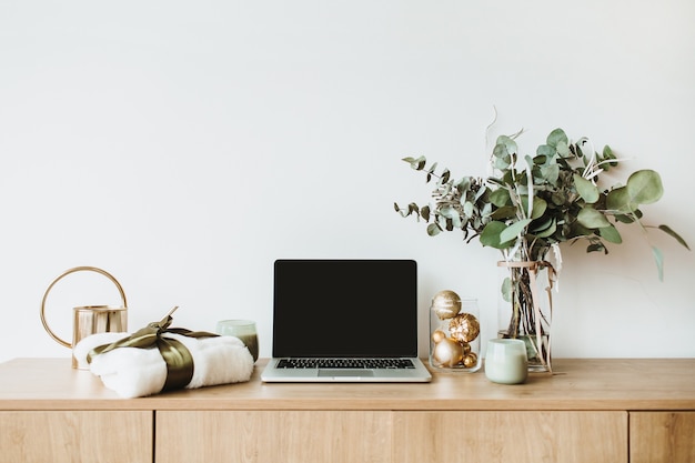 Laptop op houten bureaublad versierd met eucalyptusboeket, geschenkdoos en kaarsen op wit oppervlak