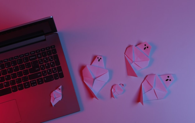 Laptop and origami ghosts in pink blue gradient neon light. Halloween theme. Top view