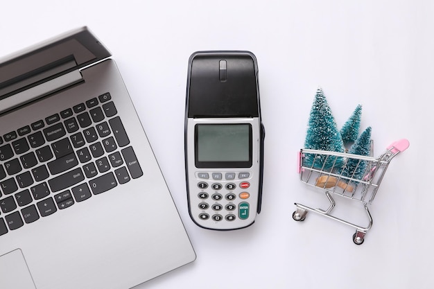 Laptop payment terminal shopping trolley with mini Christmas trees on white background Christmas shopping